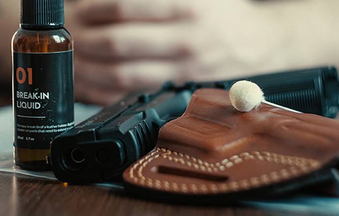 A leather OWB holster and a break-in liquid vial on a table, plastic bag underneath, a guy's arms in the background