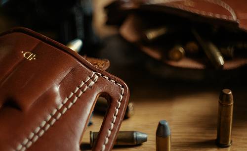 Two leather holsters lying on a table with bullets scattered around and a revolver in the background
