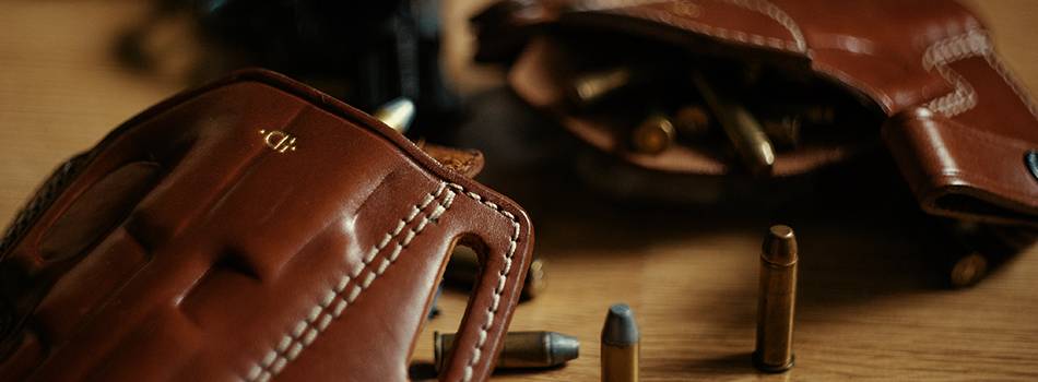 Two leather holsters lying on a table with bullets scattered around and a revolver in the background