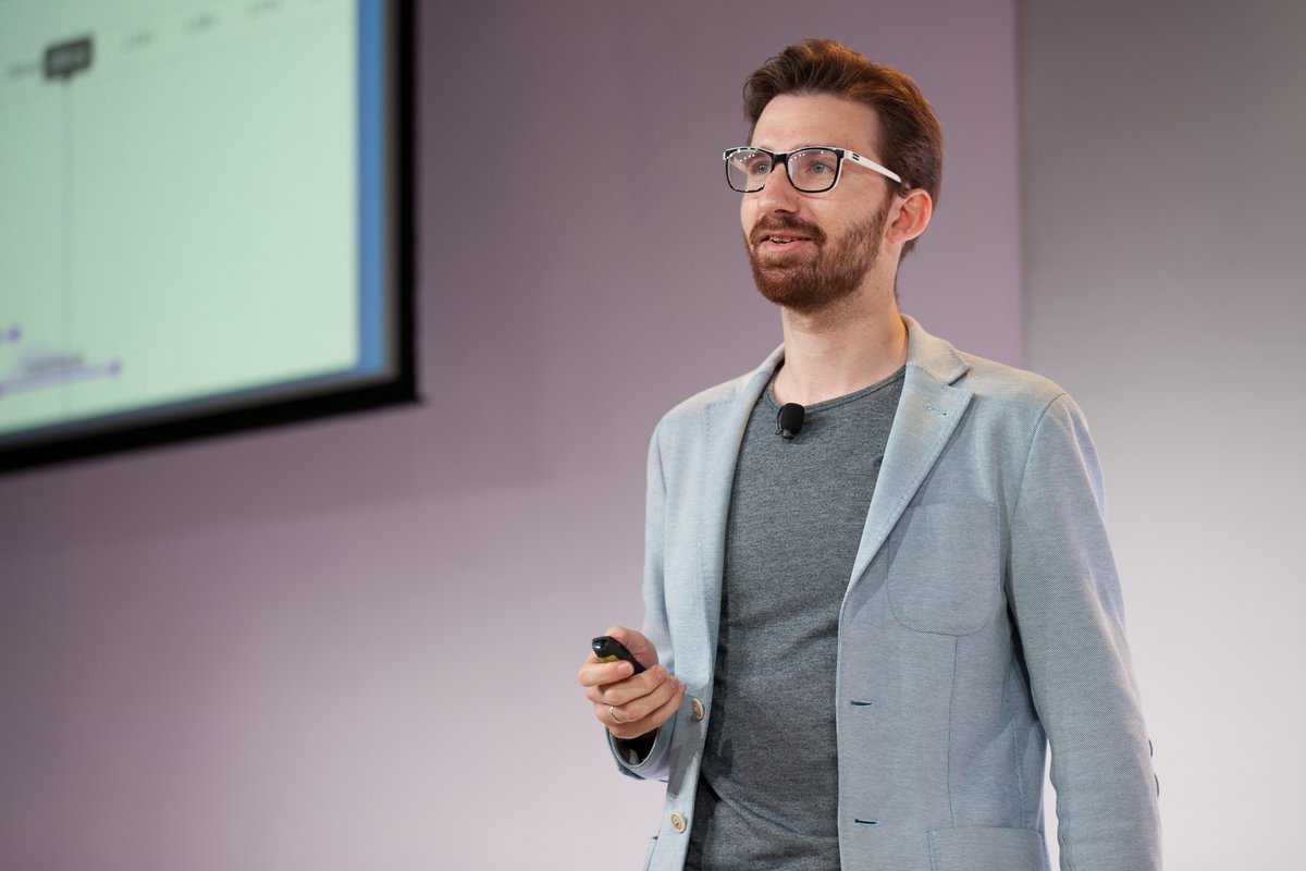 A bearded man in glasses wearing a t-shirt and blazer gives a presentation on stage.