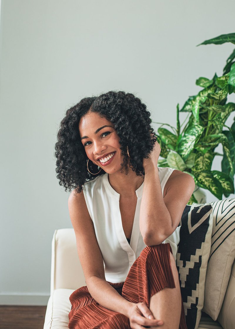 A woman with black curly hair wearing a white sleeveless blouse and red-orange striped pants sits on a white couch against patterned pillows. A tall variegated houseplant is behind her.