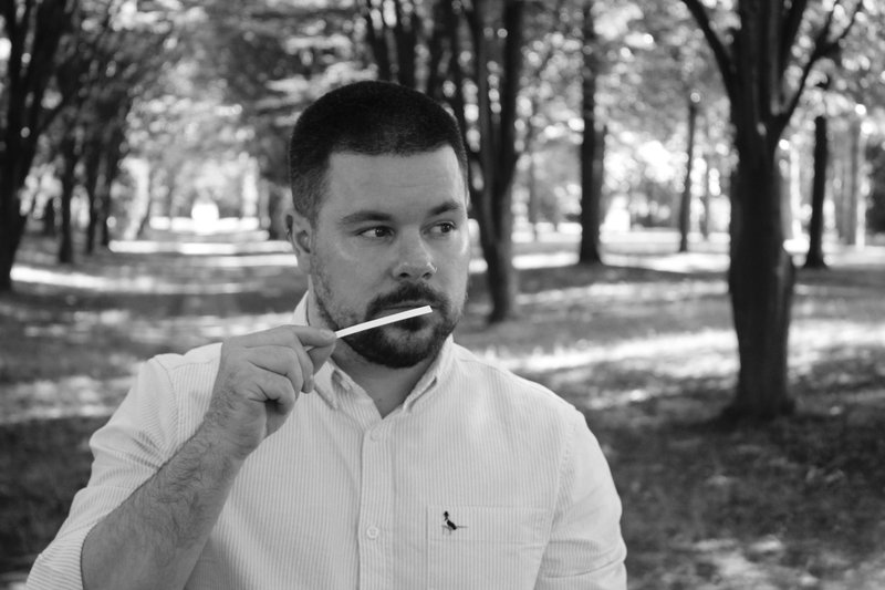 A black-and-white image of a bearded man standing in an allée of trees sniffing a fragrance sample.