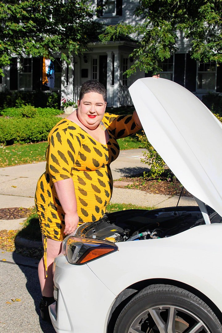 Chaya Milchtein stands at the open hood of a car. She wears a yellow patterned dress and bright red lipstick.