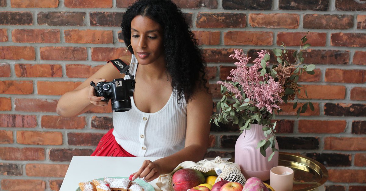 A woman sitting in front of brick wall holds a camera over a table set with food and a pink vase filled with flowers.