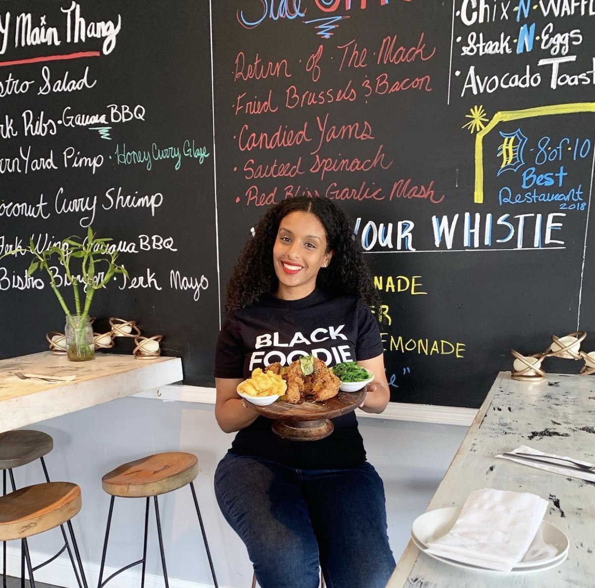 A woman with curly black hair in jeans and a black t-shirt that reads “Black Foodie” holds up a wooden platter of food in front of a chalkboard menu.