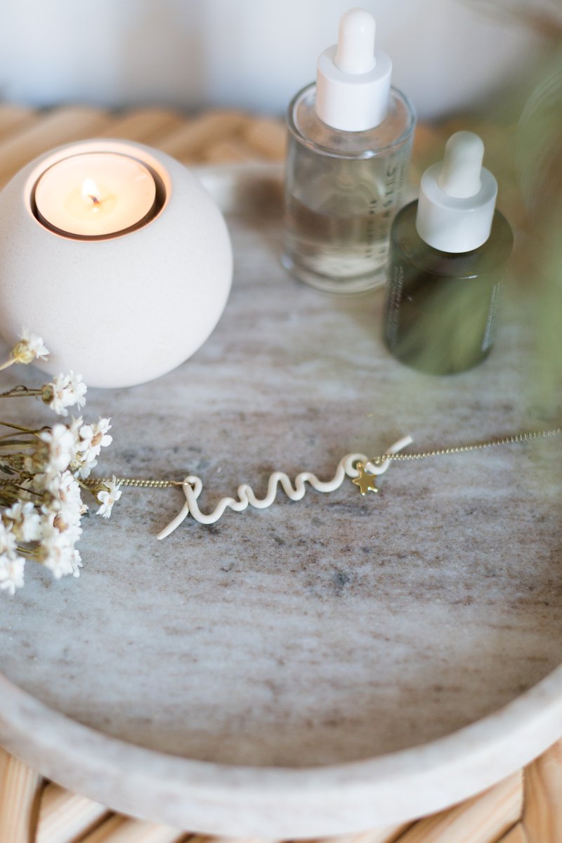 A white polymer clay necklace shaped into the name “luna” sits on a marble plate surrounded by a candle, a small bouquet of flowers and two vials.