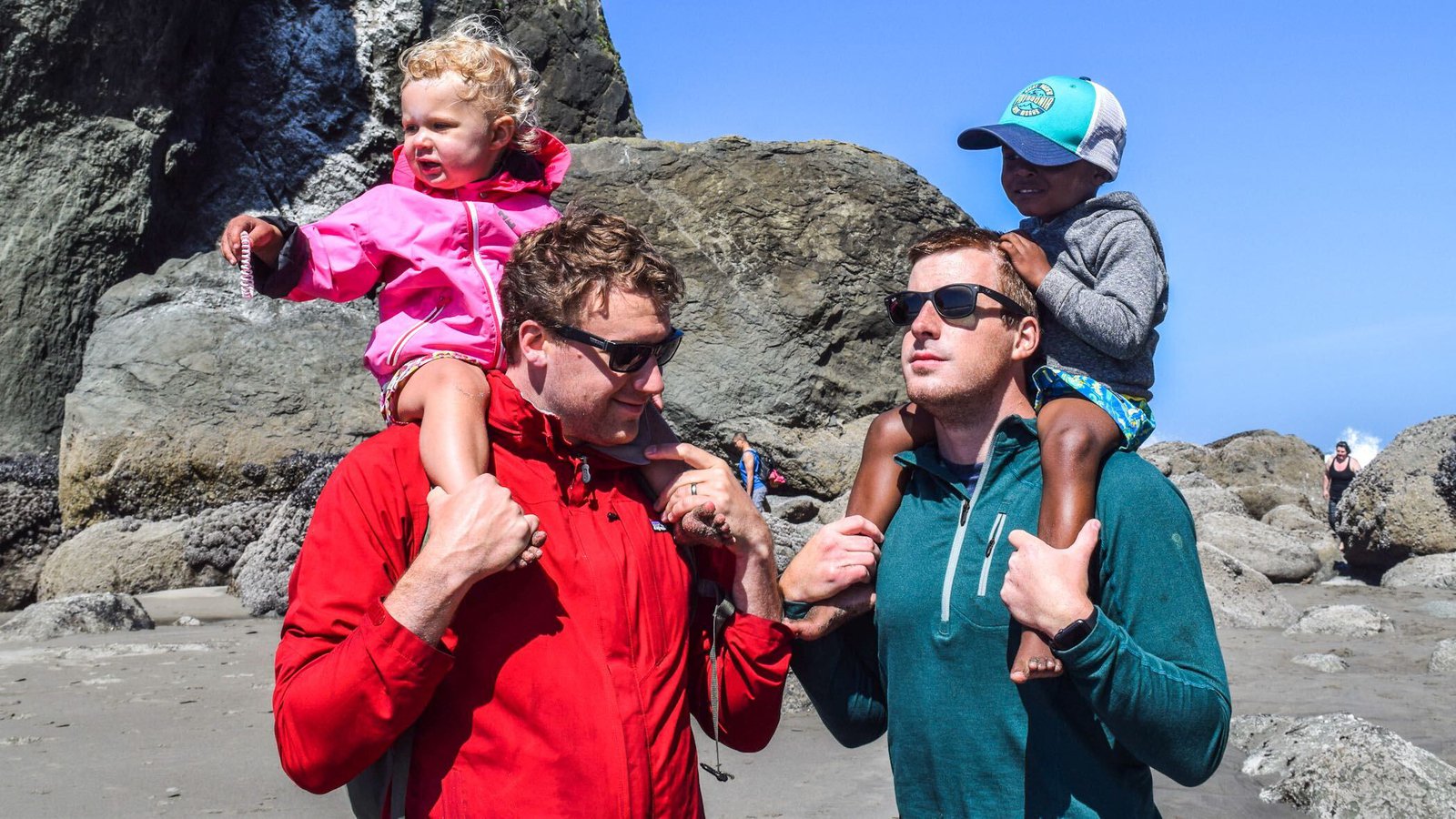 Two men in jackets and sunglasses hold toddlers on their shoulders while standing on a rocky beach.