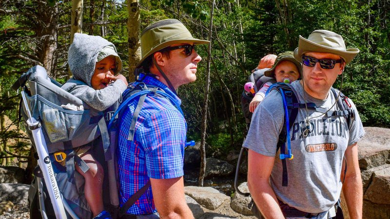 Two men wearing sunglasses and sun hats carry young children in hiking backpacks while standing in a natural setting.