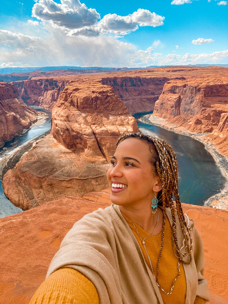 A woman in long braids wearing an orange sweater and tan cover-up stands on a plateau with a view of a water-filled canyon and a blue, cloud-filled sky behind her.