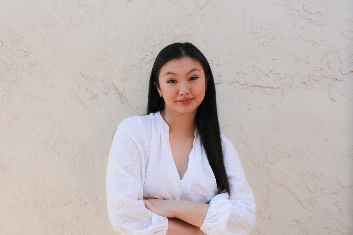 An Asian woman with long black hair wearing a white, long-sleeved blouse crosses her arms while standing in front of an off-white textured wall.