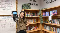 Stephanie Yi stands in her classroom in front of a bookcase with an array of colorful books. She is holding a calculator up to the camera.