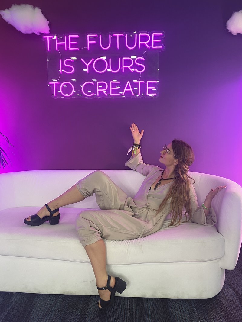 Crystal poses on a couch under a purple neon sign that reads "The future is yours to create".
