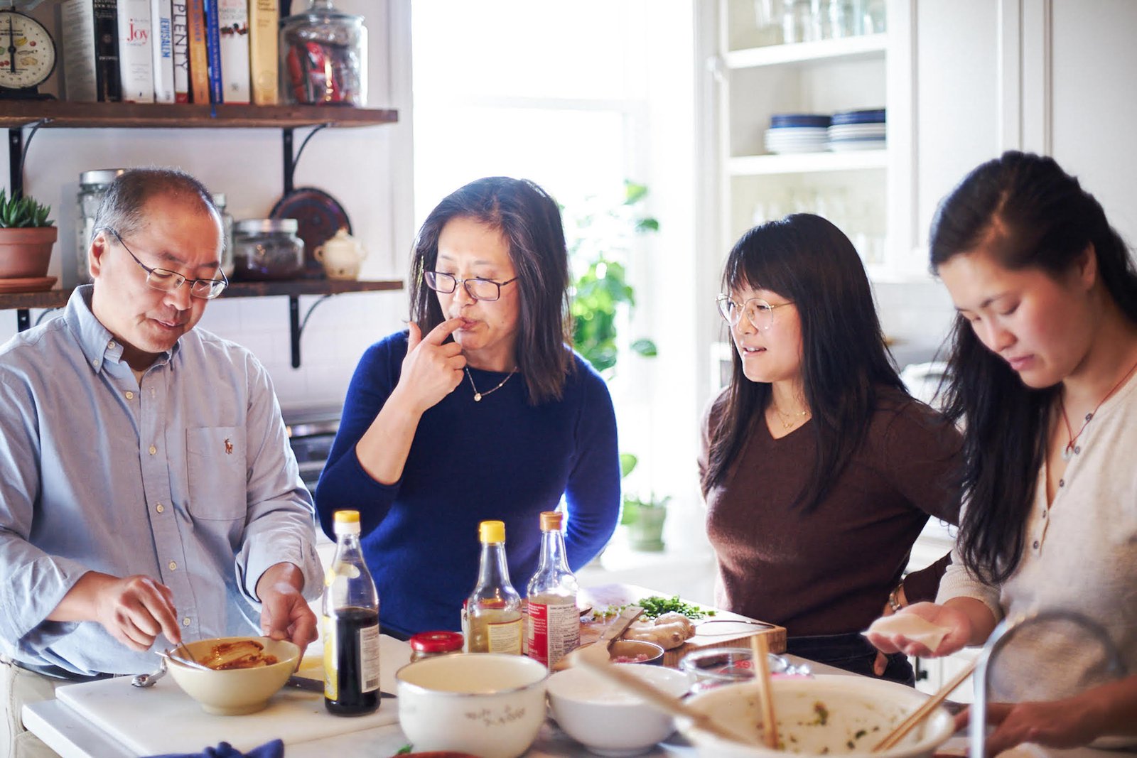 A family of four stands around a kitchen counter, preparing, considering and tasting ingredients.