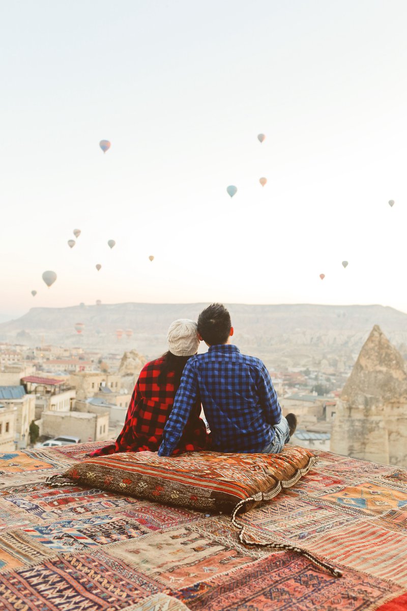 A woman and man in plaid shirts sit atop a multi-colored, patterned pillow and rugs on a lookout overseeing a view of a city below and many hot air balloons in the sky.