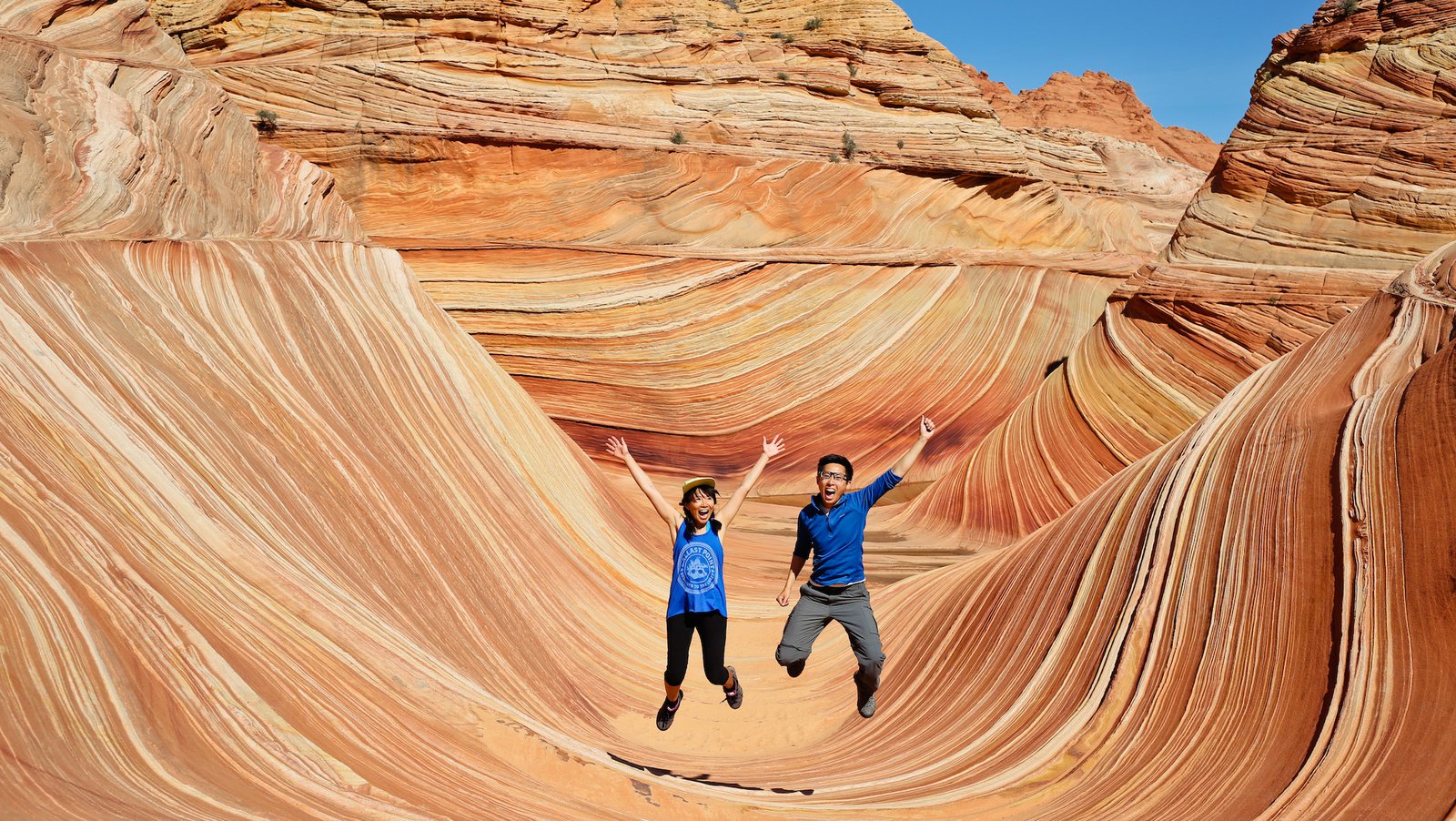 A woman and man raise their arms and open their mouths while jumping at the Wave Rock Formation in Arizona.