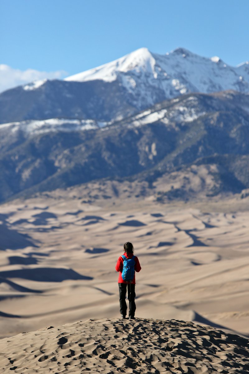 A ponytailed woman wearing a red jacket, dark pants and turquoise backpack stands on a sand dune facing a snow-capped mountain.