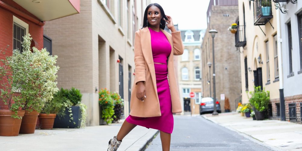 A woman in a magenta two-piece and mid-length peach coat crosses a street while looking in the distance