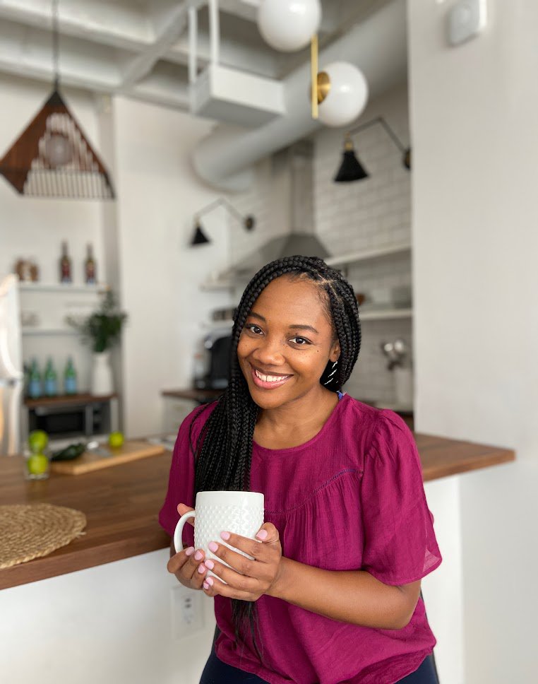 A woman with long black braids wearing a magenta top holds a large white mug in both of her hands.