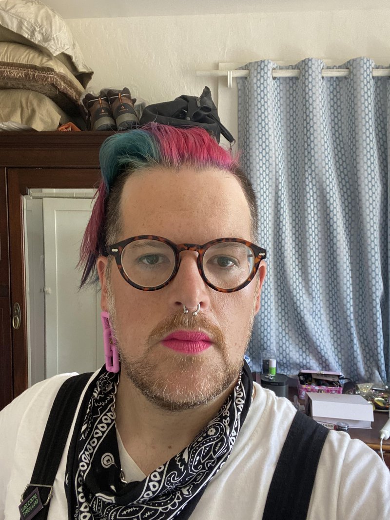 A bearded person with multi-colored hair wears glasses, a septum ring, bright pink lipstick, one dangling earring, black suspenders and a black handkerchief around their neck.