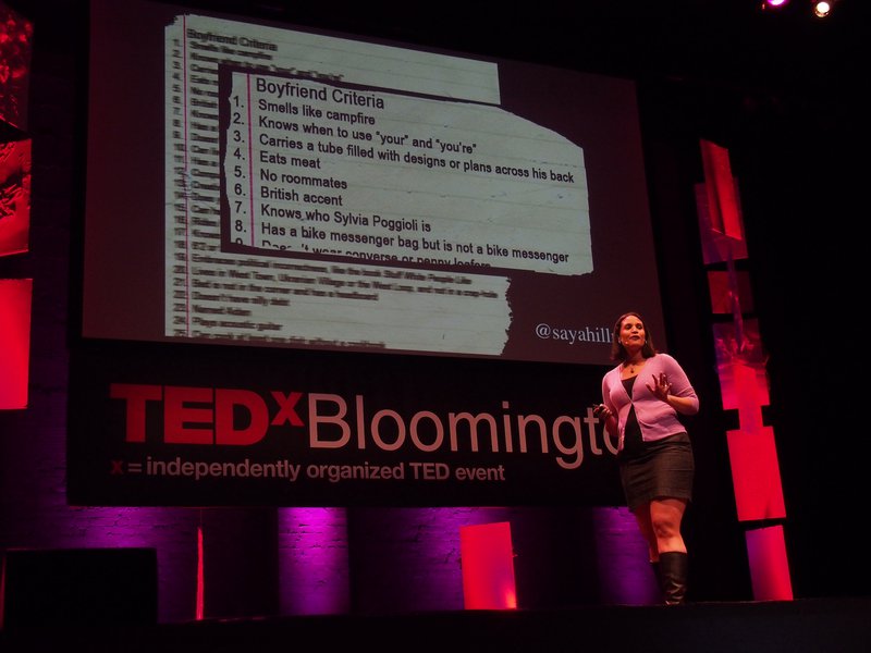 A woman wearing a pink cardigan, black dress and dark boots stands on a stage at TEDx Bloomington. A screen behind her shows a blown-up detail of a list titled “Boyfriend Criteria.”