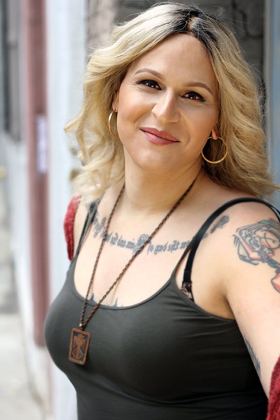 A blond woman in a black tank top with visible chest and arm tattoos poses for the camera.