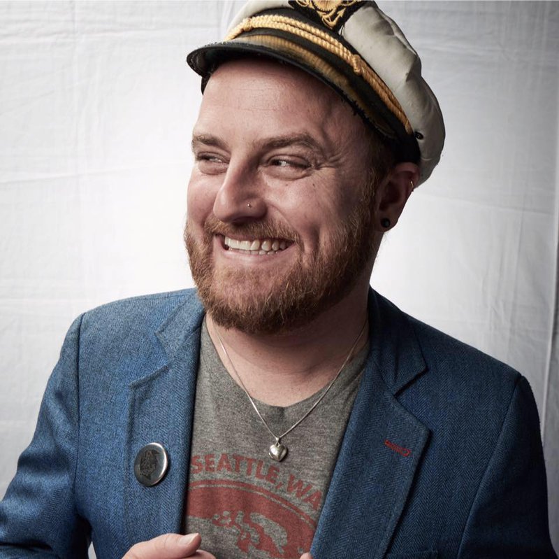 A bearded man in a blue blazer, gray t-shirt and white captain’s hat smiles and looks off to his right.