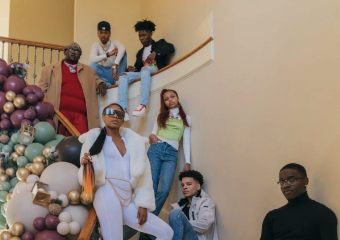 A group of fashionable, young Black people pose on a staircase surrounded by purple, green, white and gold decorations.
