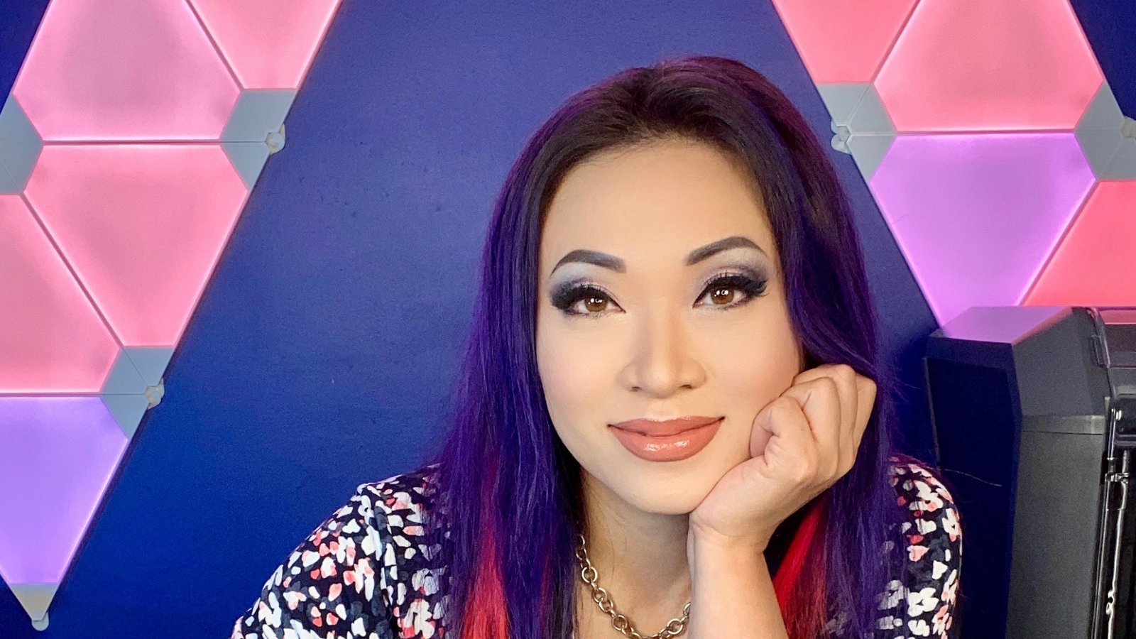 A woman with purple hair and red highlights sits with her hand against her face.