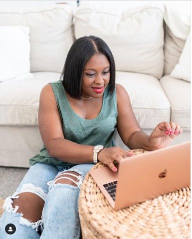 Nakisha Wynn sits on the ground with her Apple laptop propped up on a woven table. She wears ripped jeans, a t-shirt, and has a slight smile on her face.