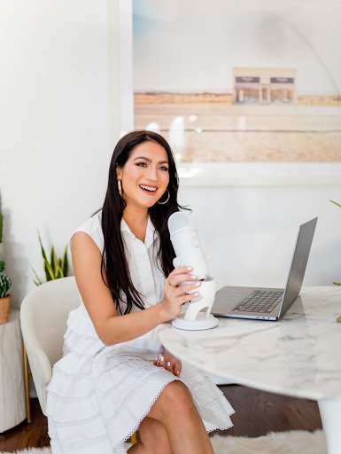 Christina Galbato sits at a marble table in front of a microphone, an open laptop at her side. She is wearing a white dress and is smiling.