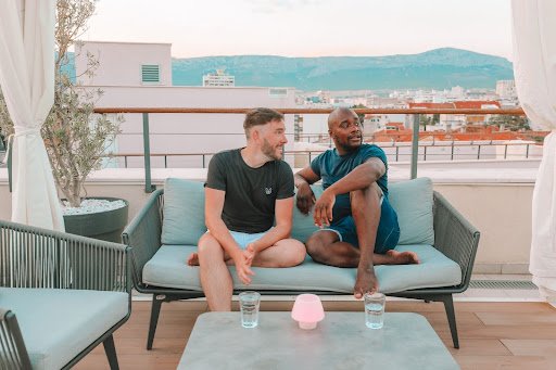 Lloyd and Yaya relax in T-shirts and shorts on a deck overlooking the city of Split, Croatia. They are seated on a green sofa with a green coffee table in the fore, with glasses of water. The cityscap