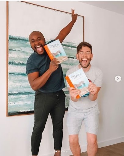 Travel bloggers Yaya and Lloyd celebrate publication of their first book. They are smiling, Yaya in black T-shirt and sweatpants and Lloyd in grey T-shirt and shorts, holding up copies of their book,
