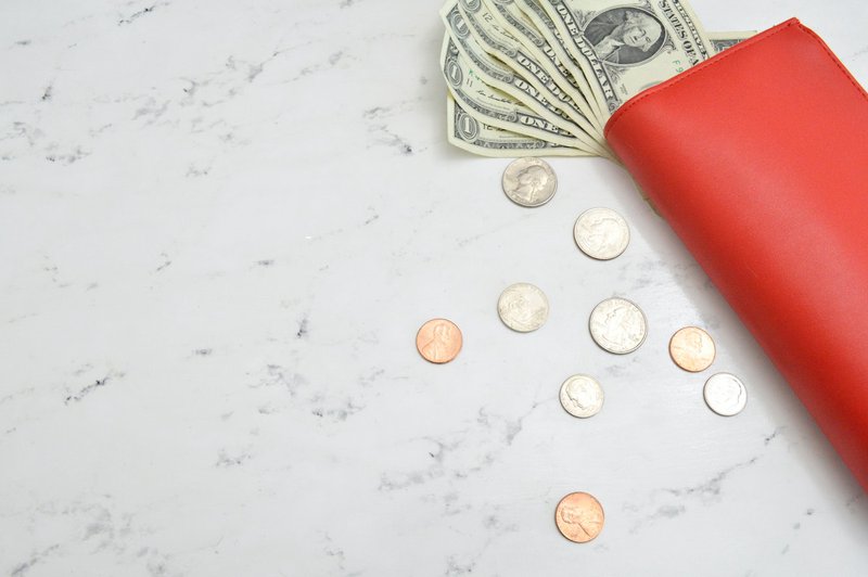Dollar bills fan out of a red wallet surrounded by coins on a marble countertop.