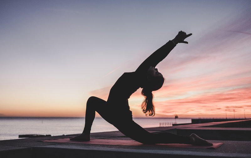 A silhouette of a ponytailed woman doing yoga on the waterfront at dusk.