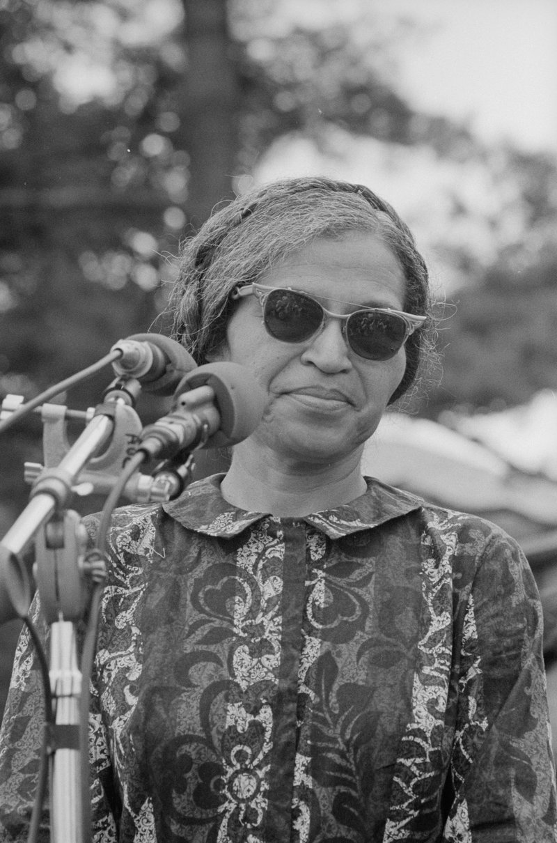 A black and white photograph of Rosa Parks, who is wearing sunglasses and a collared, patterned dress, standing outside in front of two microphones.