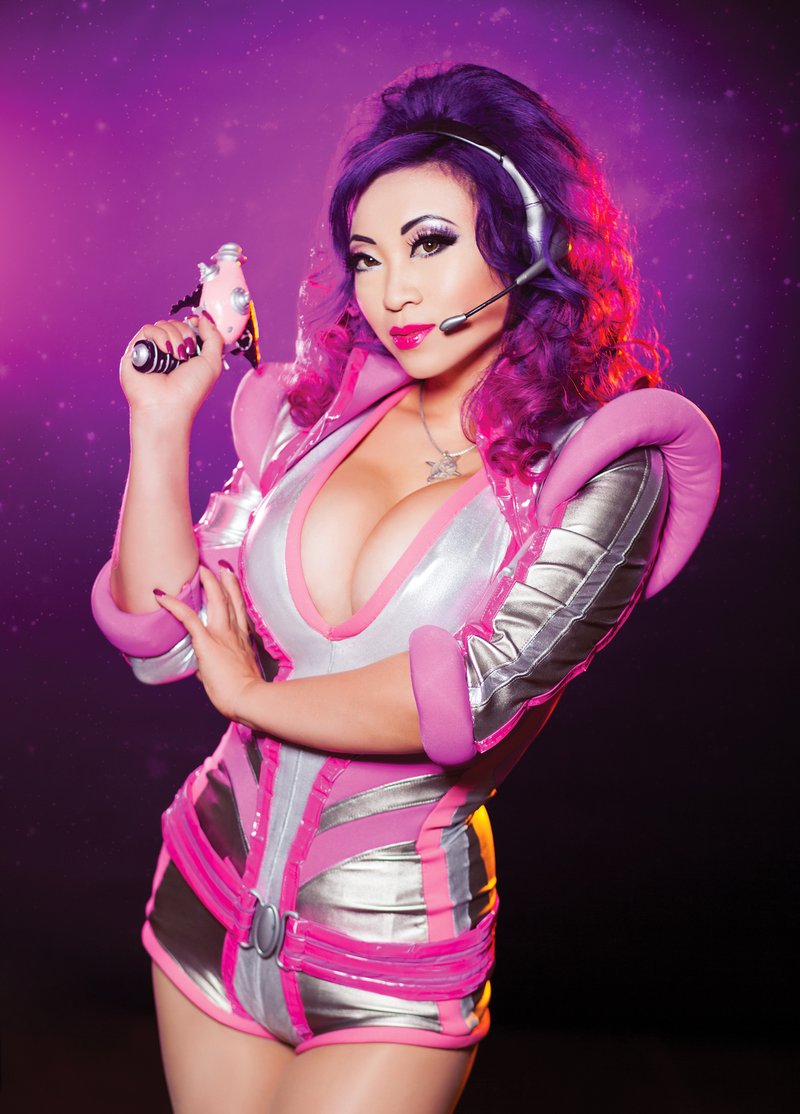 Alt-text: A woman wearing a headset and a revealing silver and pink space-themed jumpsuit holds a pink ray gun.