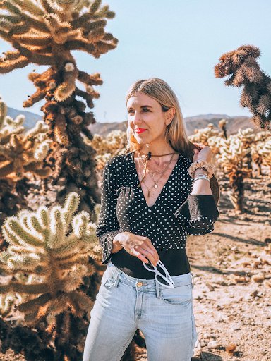Andi Eaton smiles, standing in the desert, with tall cactus plants in back of her. She wears her long blonde hair down, red lipstick, a black-and-white polka dot top and blue jeans.