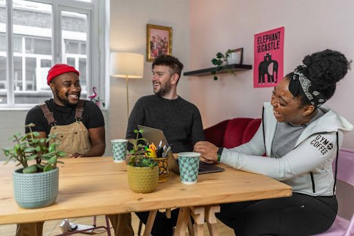 Travel bloggers Yaya and Lloyd sit at a wooden table with fitness entrepreneur Elle Linton in her bright, white-walled apartment. The group is smiling, laughing and drinking coffee from blue paper cup