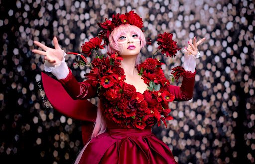 Cosplay designer Yaya Han as anime character Belle in a long-sleeved burgundy gown with red flowers on the bodice and shoulders. She wears a pink wig with a crown of red flowers and pink lipstick.