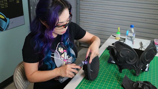 Yaya sits in her Atlanta wordshop sanding a cosplay accessory piece. She is wearing a black T-shirt with her Space Girl logo  on the front. Her hair is purple and she wears protective glasses.