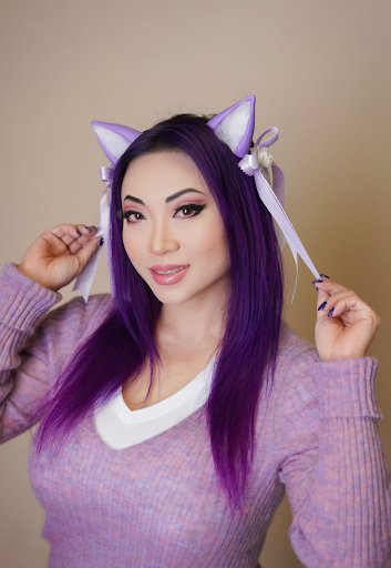 Yaya smiles, wears a lavender sweater with lavender cat’s ears embellished with ribbons. She has long purple hair and pink lipstick.