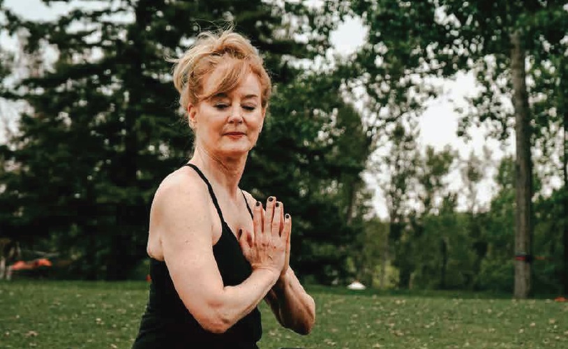 A senior woman practices yoga outside amidst a line of trees.