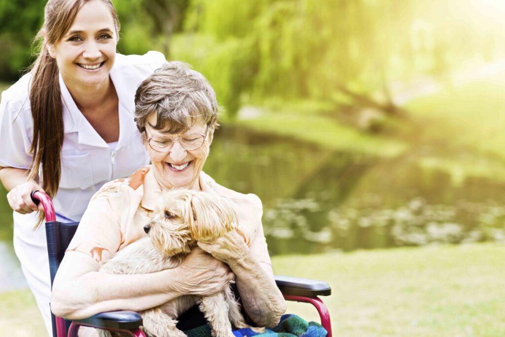 A staff member pushes a senior woman in a wheelchair holding a small dog on her lap.