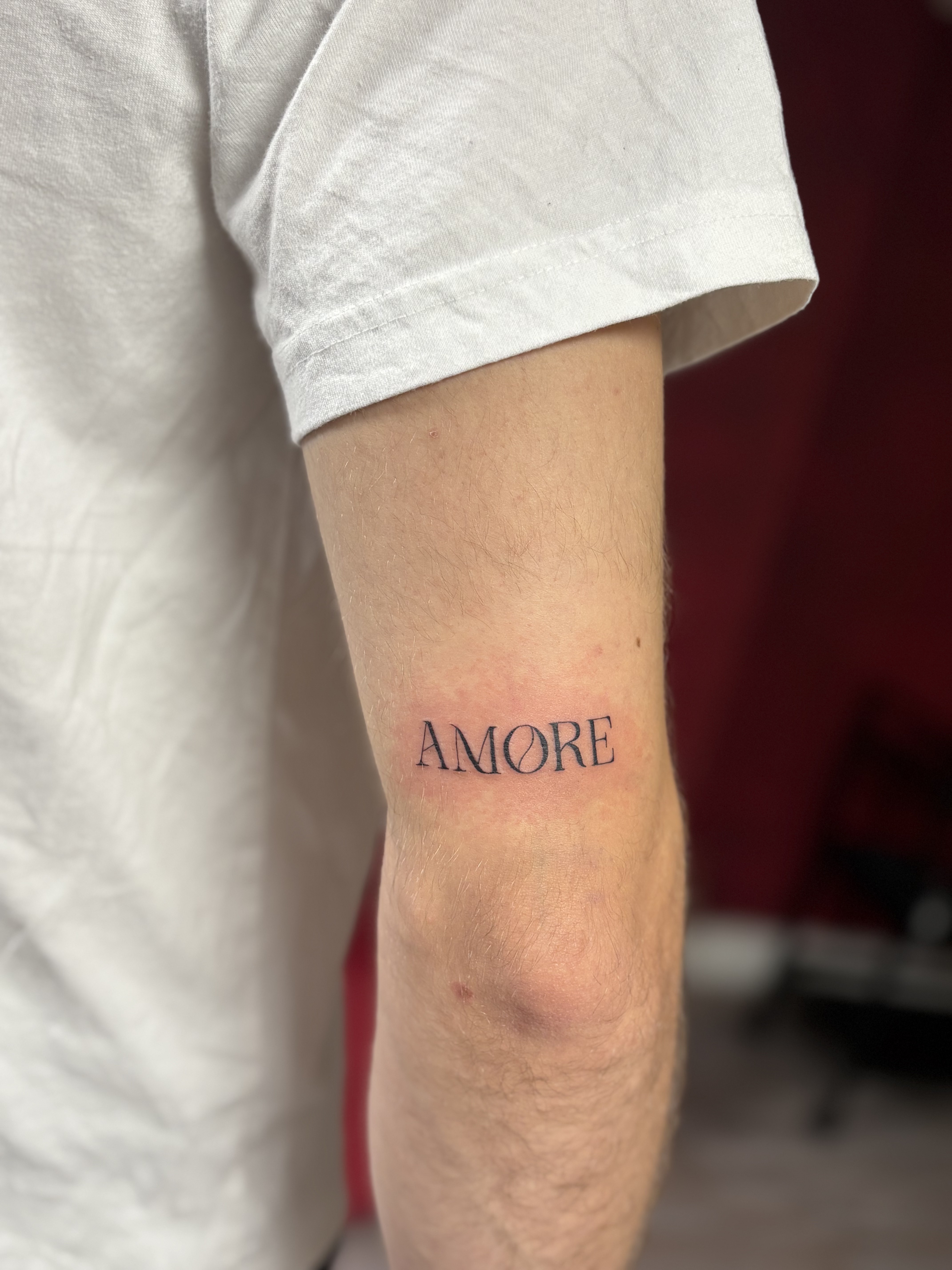 Ce tatouage présente le mot 'AMORE' réalisé dans un style lettering élégant et minimaliste. Les lettres sont exécutées en…