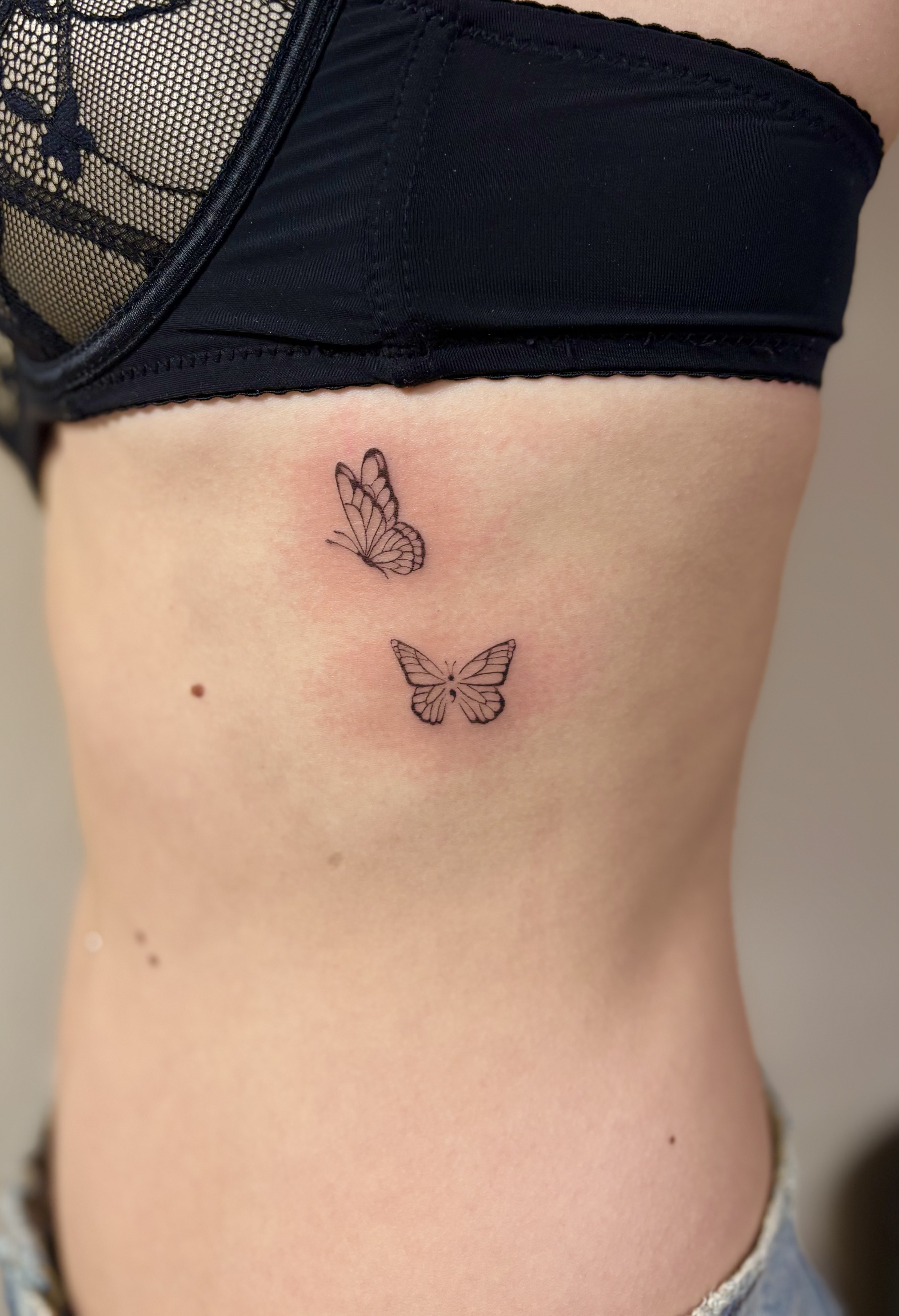 Ce tatouage présente deux papillons délicats réalisés dans un style fine-line très raffiné, positionnés sur les côtes sous…