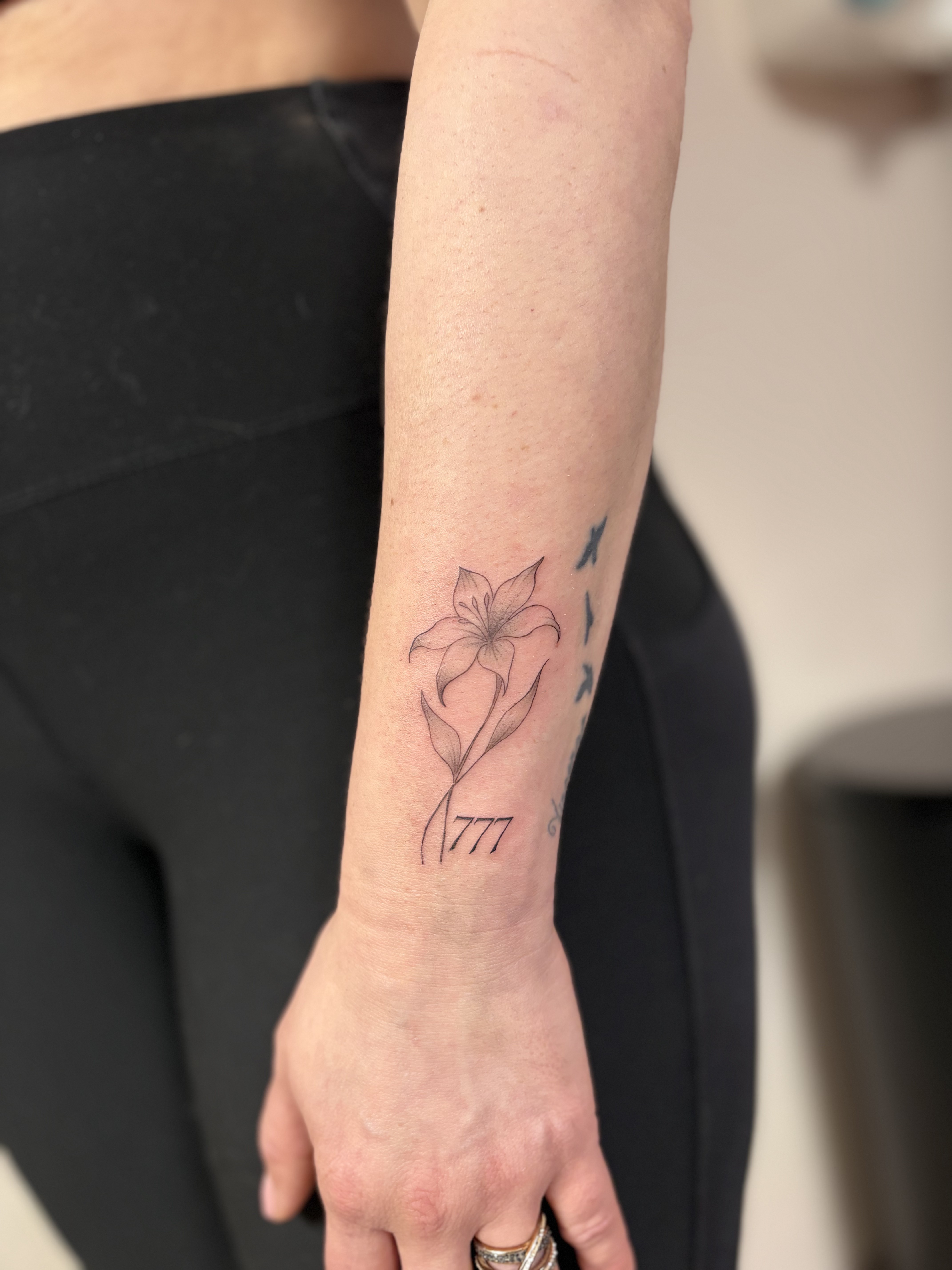 Ce tatouage présente un style fine-line délicat situé sur l'avant-bras. Le motif principal est une fleur de lys stylisée aux…