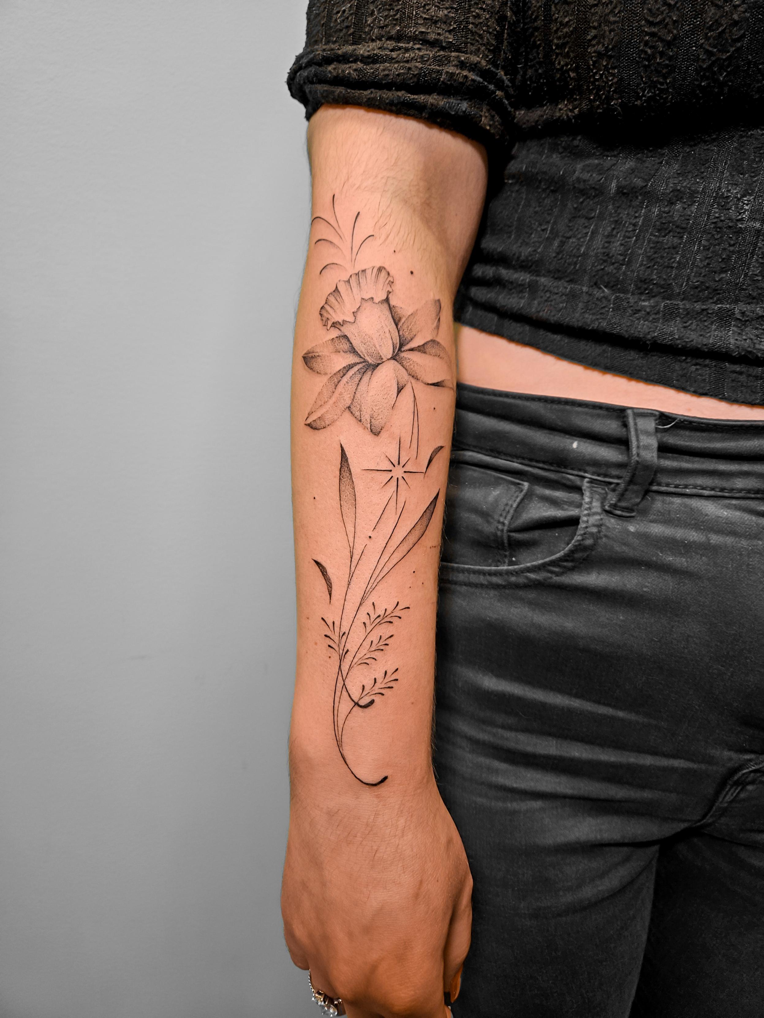 Ce tatouage floral en fine-line orne l'avant-bras dans un style délicat et élégant. Le motif principal est composé d'une…