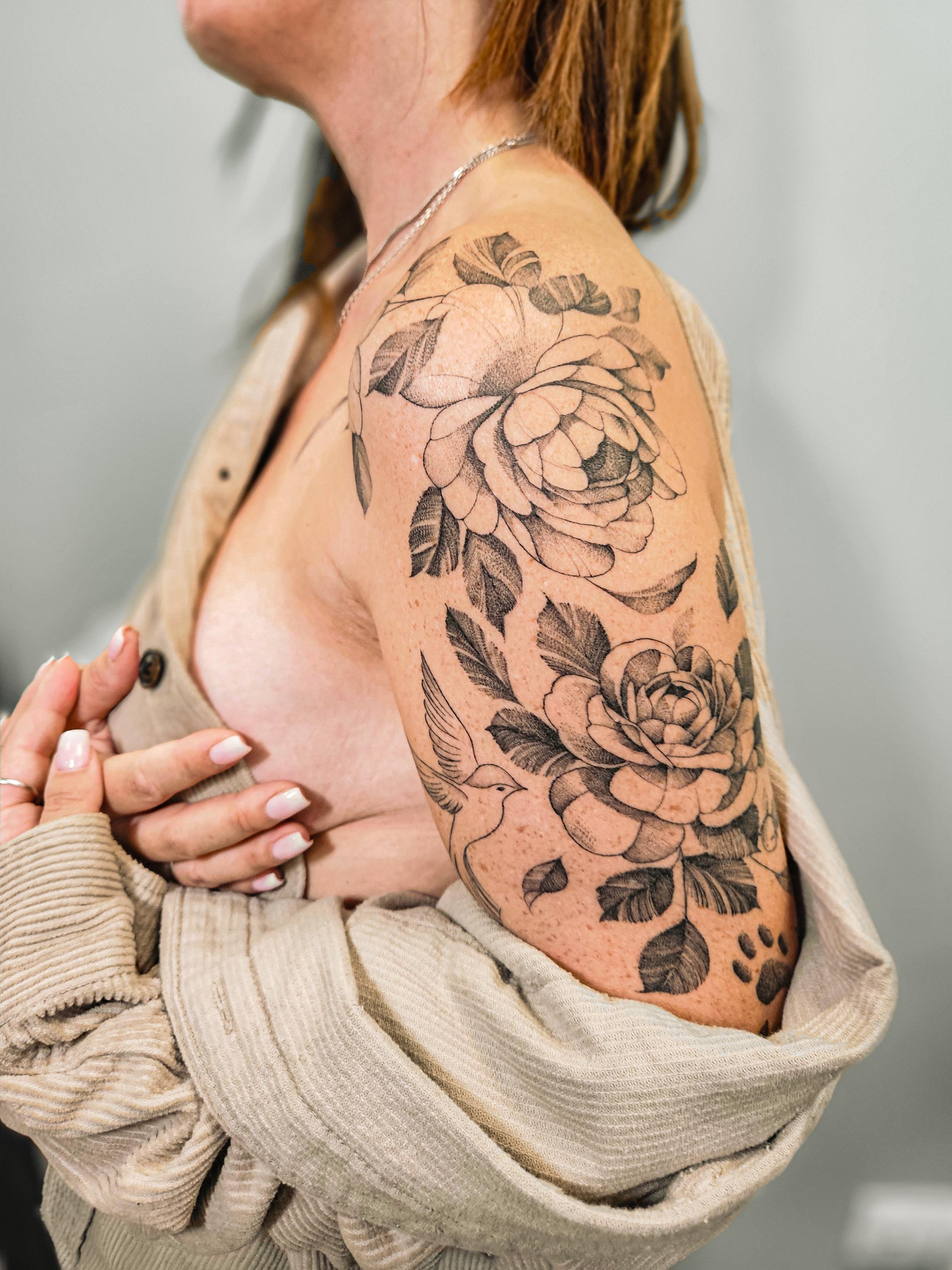 Ce magnifique tatouage floral occupe l'épaule et le haut du bras, présentant deux roses principales accompagnées de…