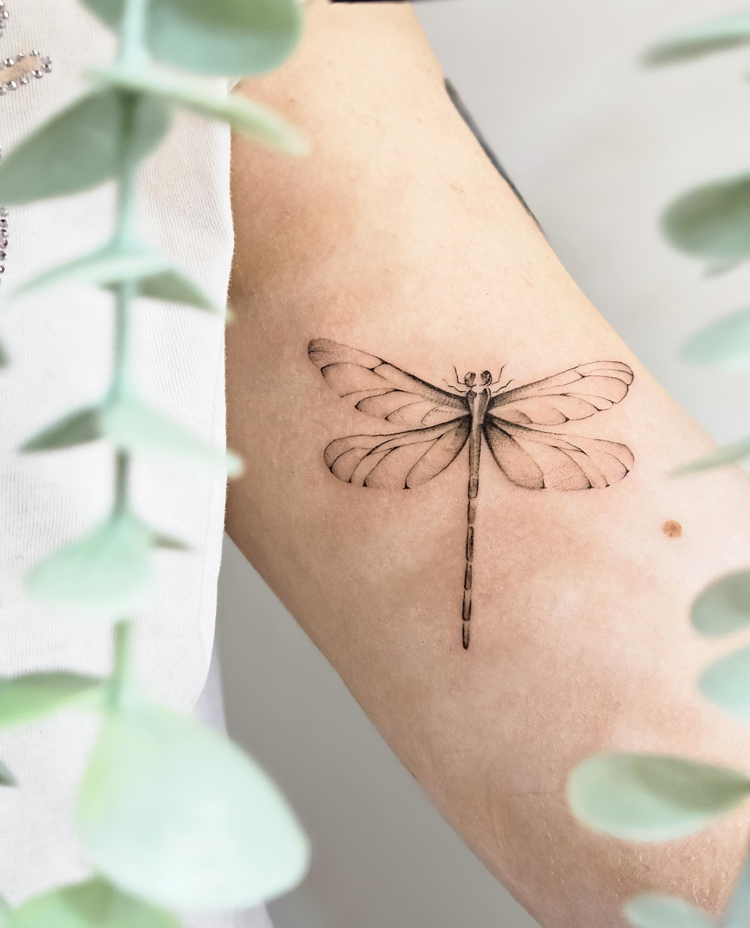 Ce tatouage représente une libellule délicatement réalisée dans un style fine-line sur l'avant-bras. L'insecte est dessiné…
