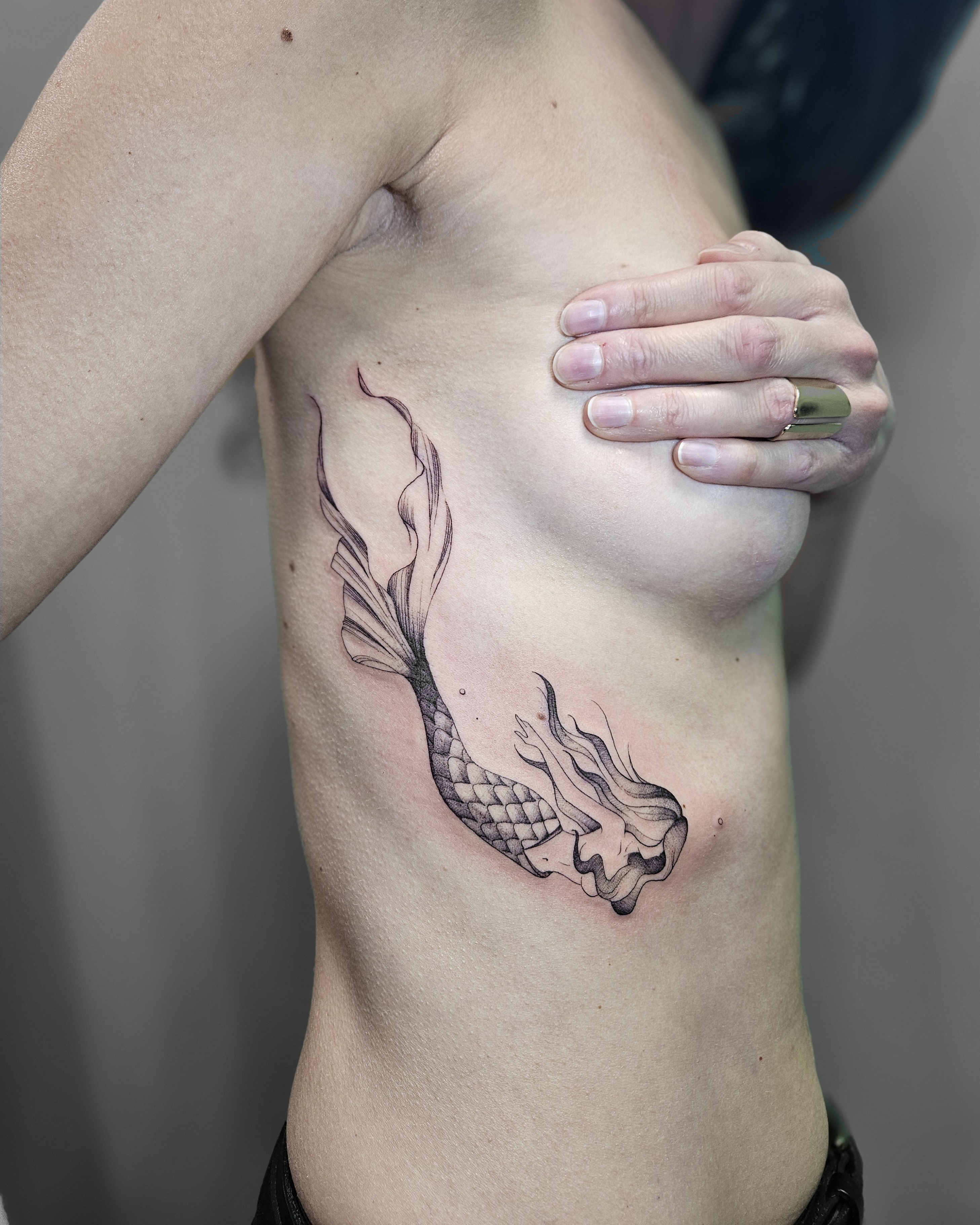 Ce tatouage représente une carpe koï stylisée dans un style fine-line avec des influences japonaises, situé sur les côtes…